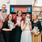 7 people standing in front of walls of books, smiling into the camera, holding pamphlets