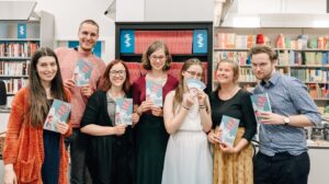 7 people standing in front of walls of books, smiling into the camera, holding pamphlets