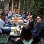 A group of people sitting at a table, enjoying drinks at a patio, books and notebooks are on the large table.