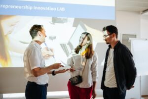 Vice-Rector Ronald Maier, left, awarding the price of the Pitch Day to Kristina and Tim. All are smiling and talking to each other.