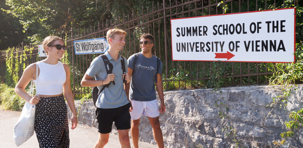 3 Students are walking and talking, behind them is a sign pointing them to a summer school