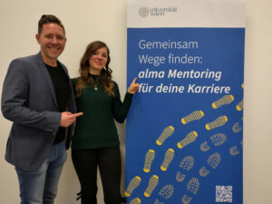 Mentee Michael und Mentorin Alexandra stehen neben einem Roll-Up des alma Mentoring Programms, beide lächeln in die Kamera und zeigen auf das Roll-Up